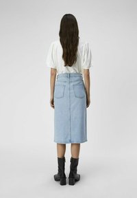 Jupe midi en denim bleu clair avec poches arrière et coupe droite, associée à des bottes noires jusqu'au genou, portée avec un chemisier blanc.