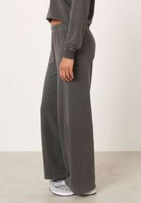 Asmuo, dėvintis tamsiai pilkas plačias kelnes, pilką ilgomis rankovėmis trumpą viršutinę dalį ir baltai pilkus sportinius batelius, stovintis ant šviesios grindų.