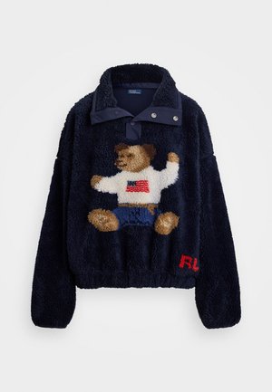 Pull à col snap en polaire marine, avec un design de ourson portant une chemise blanche et un short bleu ; accent logo rouge à l'ourlet.