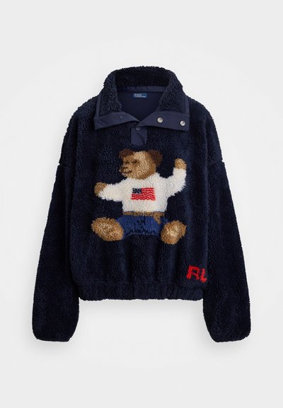 Marineblauer Fleece-Pullover mit einem Druckknopfkragen, der ein Design eines Teddybären zeigt, der ein weißes Hemd und blaue Shorts trägt; rotes Logo-Detail am Saum.