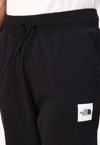 Sorte sweatpants lavet af en blanding af bomuldsstof, med en ribbet talje, snoretræk og et hvidt logo patch på siden.