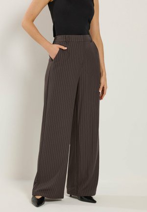 Femme portant un pantalon large à rayures fines marron foncé, un haut sans manches noir et des chaussures noires pointues, debout avec une main dans la poche.
