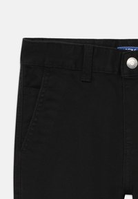 Nærbilde av svarte denimbukser som viser midjebånd, beltehempe, sølvknapp og detaljer av frontlommen på hvit bakgrunn.