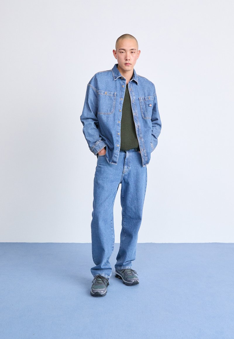 Chaqueta de mezclilla con dos bolsillos en el pecho, combinada con jeans azules a juego y zapatillas deportivas grises. La capa interior es un suéter de punto verde oscuro.