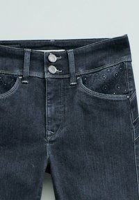 Pantalones vaqueros de mezclilla oscura con doble botonadura plateada, costuras blancas, trabillas para cinturón y detalles de tela estampada en los bolsillos delanteros.