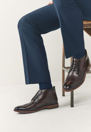 Brune læderankelstøvler med brogue-detaljer og rundt tådesign. Kombineret med mørkeblå skræddersyede bukser og marineblå sokker.