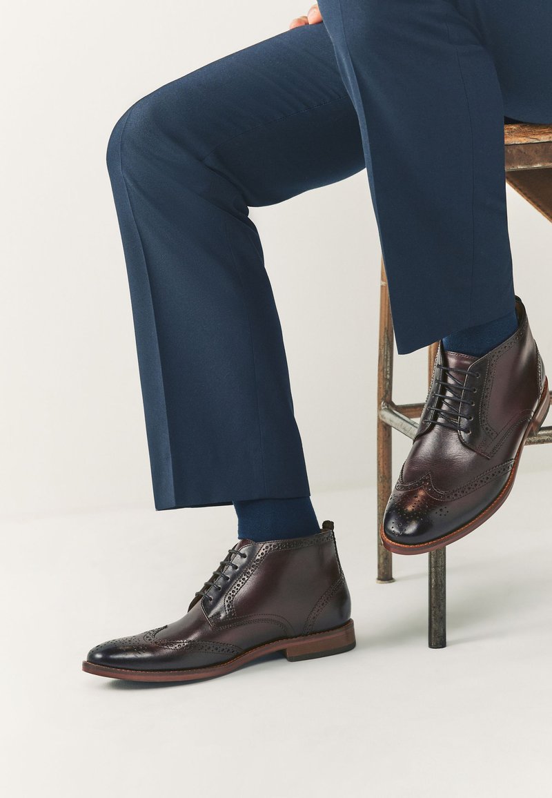 Braune Leder-Stiefeletten mit Brogue-Details und runder Zehenpartie. Kombiniert mit dunkelblauen, maßgeschneiderten Hosen und marineblauen Socken.