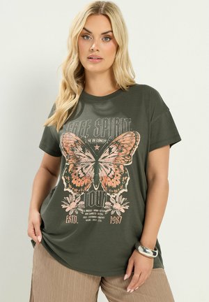 Blonde woman wearing an olive green T-shirt with a large pink butterfly graphic and beige pants, standing against a plain background.