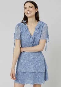 Robe fleurie bleue avec encolure à volants, jupe superposée et courtes manches transparentes liées aux poignets. Fabriquée en tissu léger et texturé.