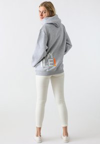 Felpa grigia con cappuccio e grande grafica "SMILE!" bianca e arancione sulla schiena, abbinata a pantaloni bianchi slim-fit e calzature chiare.