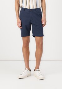 Navy blue shorts made of cotton blend, featuring a button closure, flat front design, and rolled hems, paired with white sneakers.