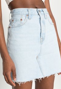 Mörkhyad person iklädd en blå denim mini kjol med fransar och en vit kort topp mot en neutral bakgrund.