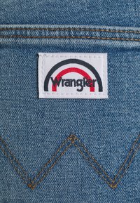 Wrangler denim-märke har en vit etikett med en regnbågsdesign i röd, svart och grå. Texturerat blått tyg med kontrasterande gula sömmar.