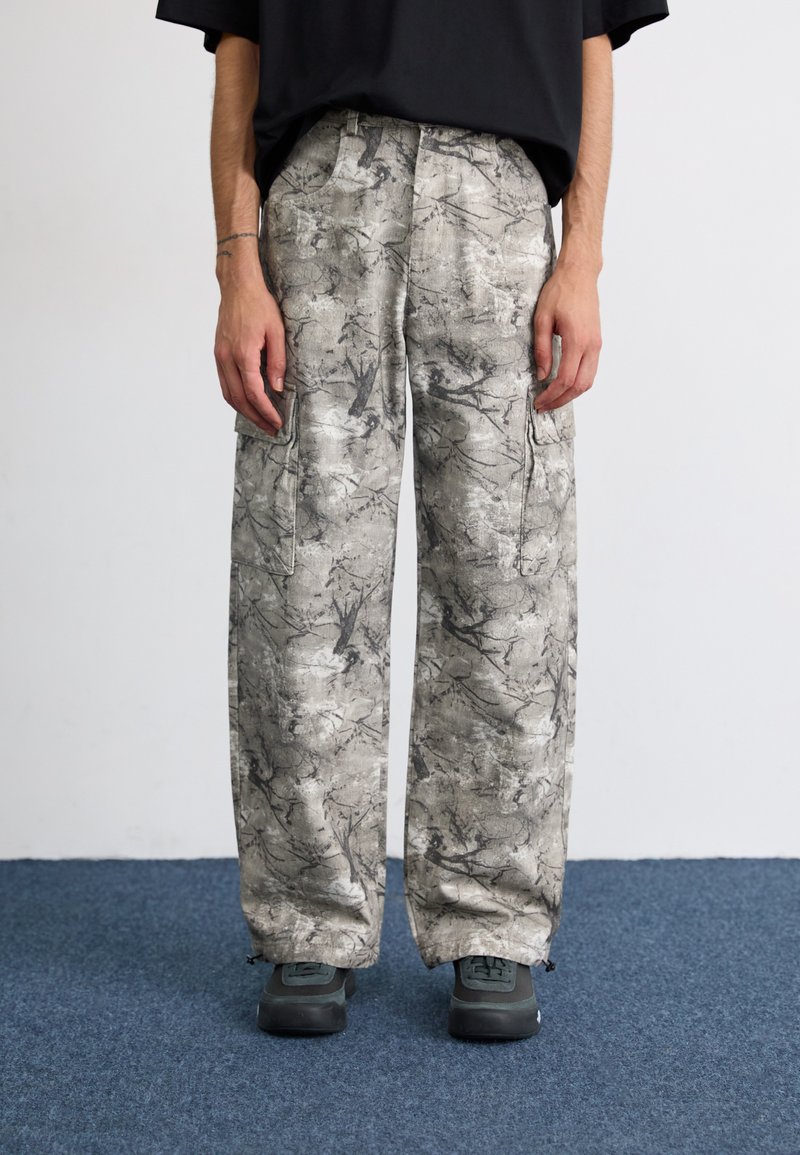 Pantalones cargo de pierna ancha en una tela gris texturizada con patrones abstractos en blanco y negro, que cuentan con bolsillos laterales y puños con cremallera.
