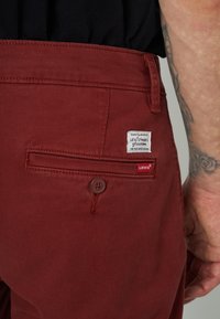 Pantaloni bordeaux con una texture liscia, dotati di una tasca sul retro e un'etichetta bianca Levi's. Accent di bottone marrone vicino alla tasca.