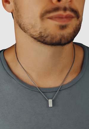 Silbernes rechteckiges Anhänger-Neklace an einer metallischen Kugelkette, getragen über einem grauen Rundhalsausschnitt-Shirt. Einfaches Design mit glatter Oberfläche.