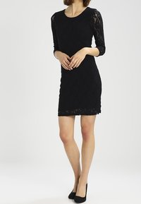 Robe en dentelle noire avec manches trois-quarts, silhouette ajustée, ourlet festonné et motif floral. Associée à des chaussures à talons noires.