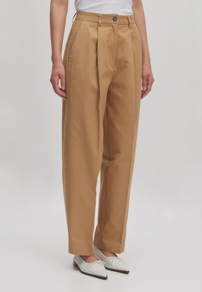 Beige, wijde broek met plooien aan de voorkant, een rechte pasvorm, hoge taille en een enkele knoopsluiting. Gecombineerd met witte puntige schoenen.
