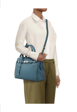 Person trägt ein weißes Hemd mit Knöpfen und olivfarbene Hose und hält eine blaue Lederhandtasche mit oberen Griffen und einem Schulterriemen.