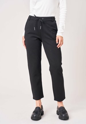 Persoon met zwarte cropped broek met aantrekkoord in de taille, zwarte loafers en een wit geribd shirt met lange mouwen, staand tegen een effen lichte achtergrond.