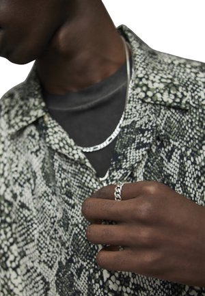 Camicia a fantasia serpente verde e bianca sopra una maglietta nera, collana in argento, e anello intrecciato in argento su una mano. Texture morbide, motivi intricati.