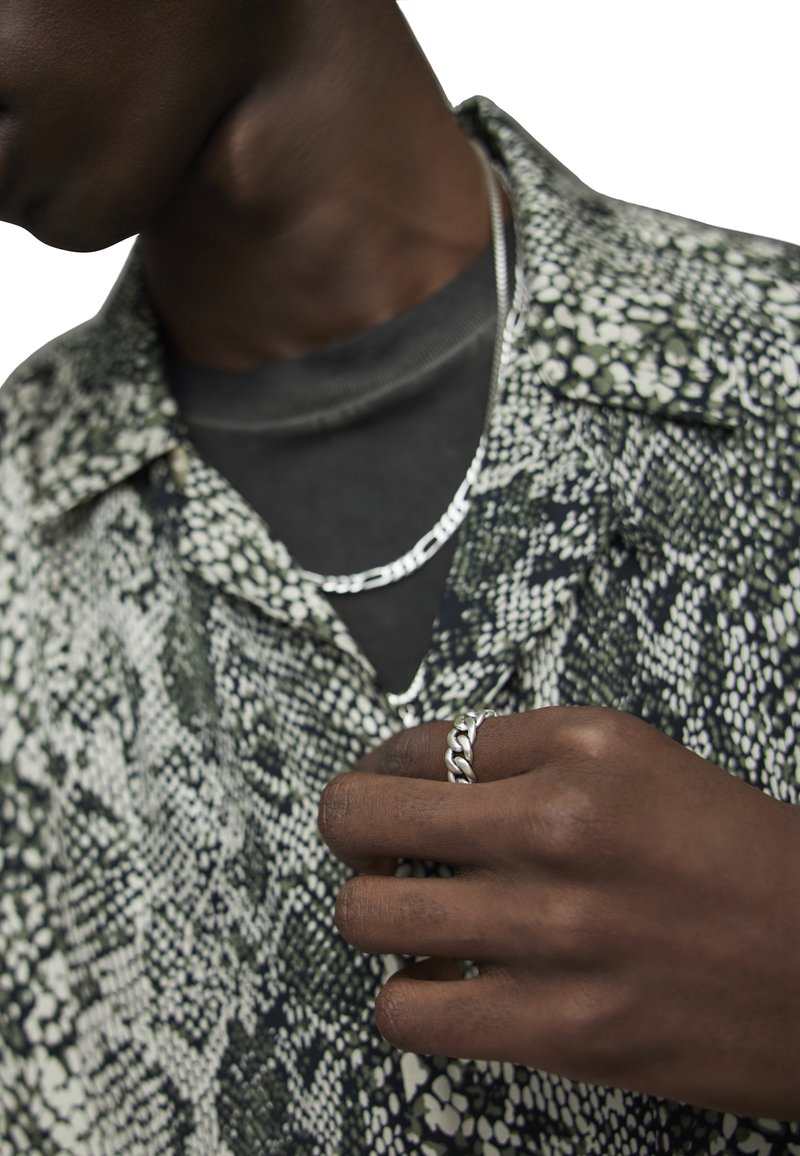 Chemise à imprimé serpent vert et blanc par-dessus un top noir, collier en chaîne argentée et bague en argent tressée sur une main. Textures lisses, motifs complexes.
