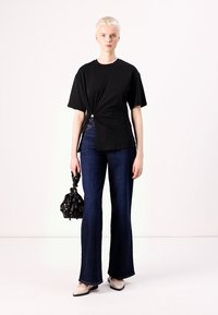 Tricou negru legat, blugi întunecați evazați și balerini bej. Accesoriu: geantă mică neagră cu model decorativ. Design simplu și modern.