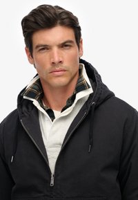 Zwarte hoodie-jacket met een ritssluiting over een wit poloshirt. De jas heeft een gewatteerde voering en een geruit kraag in donkergroen en beige.