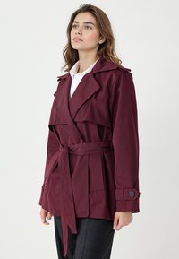 Bordowy płaszcz trencz z szerokim kołnierzem, paskiem w talii i zapinanymi na guziki mankietami. Gładka tkanina o dopasowanym kroju, z przednimi kieszeniami.