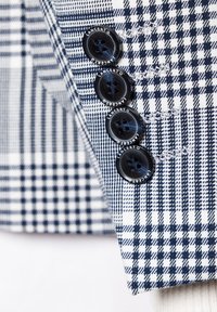 Materiał w granatowo-białą kratkę z czterema granatowymi guzikami z logo oraz widocznymi detalami szycia na rękawie dopasowanej marynarki.