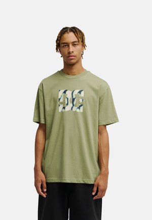 Młody mężczyzna z krótkimi warkoczykami ma na sobie luźny oliwkowozielony t-shirt z dużym wzorzystym logo „DC” na piersi, stojąc na jednolitym tle.