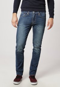 Homme portant un jean slim bleu, un pull bleu marine foncé et des baskets bordeaux, debout devant un fond clair uni.