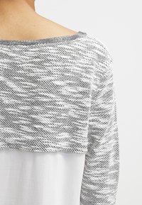 Grau-weißes, strukturiertes Strickoberteil mit weitem Ausschnitt und einem geschichteten Design, das einen glatten, weißen unteren Abschnitt umfasst.