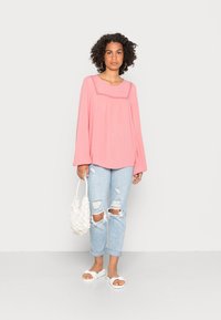 Top rosa de mangas longas com acabamento em renda, combinado com jeans claros desfiados e sandálias brancas. Modelo segurando uma bolsa de palha branca.