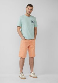 T-shirt en coton bleu clair avec un motif de palmier, associé à un short pêche. Le modèle porte des baskets beiges et blanches. Tenue estivale simple et décontractée.