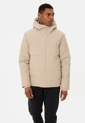 Beige isolierte Jacke mit hohem Kragen und Kapuze, mit gesteppter Oberfläche und elastischen Bündchen, getragen über dunklen Hosen.