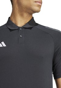 Zwarte poloshirt van een gestructureerde stof, met een klassieke kraag en twee knopen, versierd met witte logodetails op de linkerkant van de borst en de schouders.