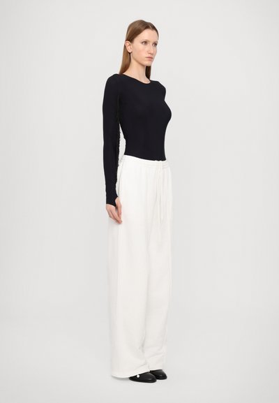 Black long-sleeve top with textured side detailing, paired with loose-fitting white trousers. Footwear is black, rounded-toe design.
