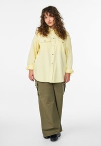 Žena s kučeravými vlasmi, oblečená v žltej košeli na gombíky, olivovo zelených nákladných nohaviciach a čiernych topánkach, stojaca pred bielym pozadím.