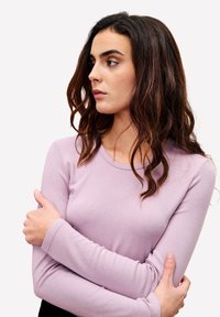 Junge Frau mit langen, welligen, braunen Haaren in einem lila Langarmshirt, mit verschränkten Armen, schaut nach links vor einem schlichten weißen Hintergrund.