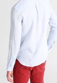 Ljusblå linneskjorta med långa ärmar, knappade manschetter och en bakficka, i kombination med röda byxor. Den texturerade tyget har ett subtilt vävmönster.