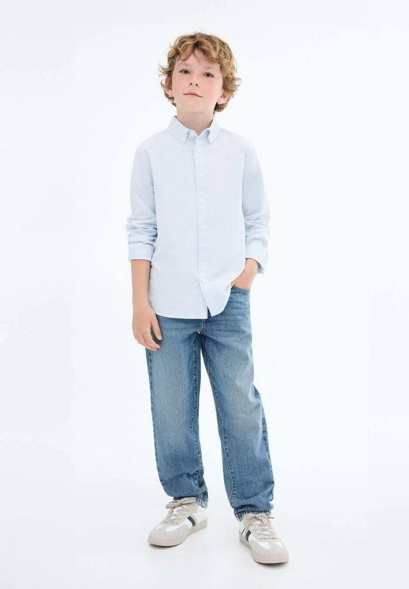 Cămașă albastră deschis cu dungi, blugi largi de culoare albastră și adidași crem cu accente bleumarin. Copilul stă cu mâinile în buzunare.