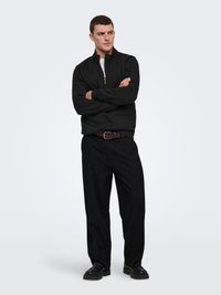 Homme debout, bras croisés, portant un pull à demi-fermeture éclair noir, un pantalon noir, une ceinture marron et des chaussures noires sur fond blanc.