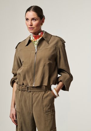 Femme aux cheveux tirés en arrière portant une tenue utilitaire marron avec manches retroussées, un foulard à fleurs et trois bracelets à son poignet, sur un fond uni.