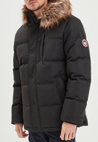 Giacca a vento nera con cappuccio in pelliccia sintetica removibile, dotata di zip frontale, bottoni a scatto e tasche laterali. Design isolato con finitura texturizzata.