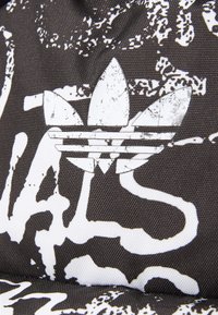 Juodas audinys su nusidėvėjusiu baltu Adidas logotipu ir abstrakčiais baltais ženklais. Paviršius yra lygus ir šiek tiek šiurkštus.