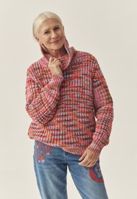 Kootud kampsun kõrge kraega mitmevärvilistes punastes, roosades ja pruunides toonides. Laisalt istuv, soonikkoes varrukapitsid. Sinised teksad lilleliste tikanditega detailid.