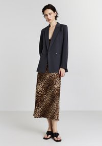 Femme portant un blazer noir sur un top noir, une jupe midi à imprimé léopard et des sandales à bout découvert noires, se tenant devant un fond gris clair uni.