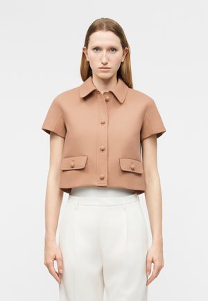 Kvinde iført en kortærmet, cropped beige jakke med knapper og forlommer, kombineret med hvide bukser med høj talje.