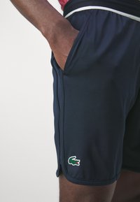 Námořnické výkonnostní šortky z lehkého materiálu mají boční kapsy a na spodním lemu zelené logo krokodýla, s bílým kontrastním šitím.